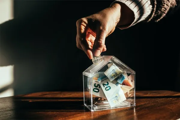 Hand wirft Geld in eine Spardose in Hausform