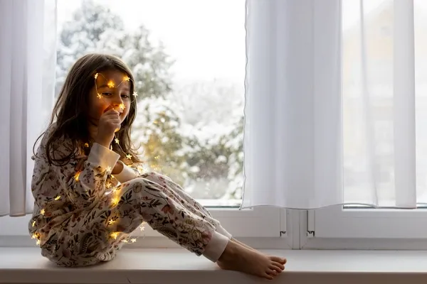 Mädchen mit Lichterkette sitzt neben Fenster